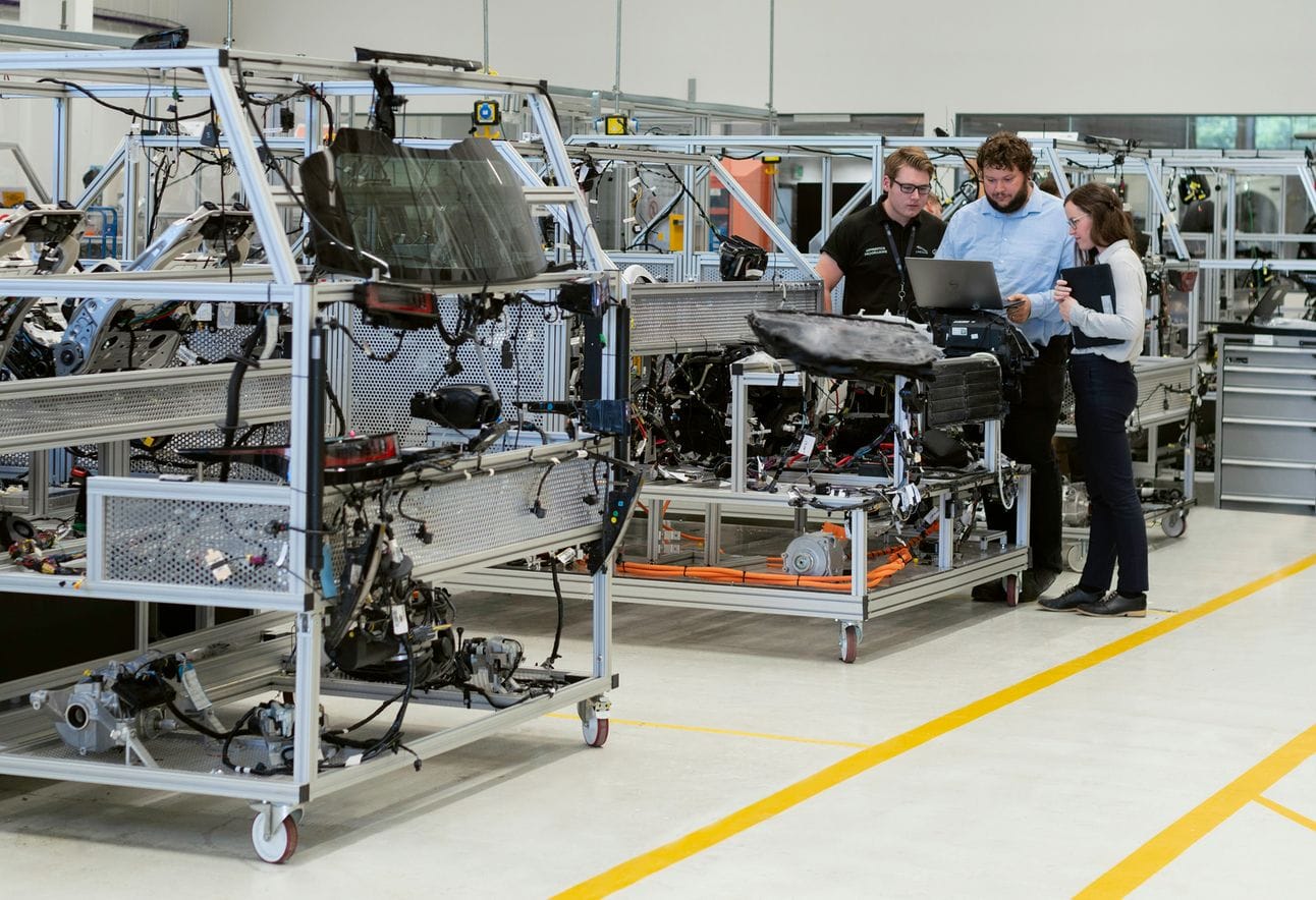 Three engineers in a factory setting are reviewing data on a laptop while standing next to a partially assembled vehicle on an industrial assembly line. The workspace is filled with metal frames, machinery, and various automotive components, indicating a high-tech manufacturing environment.