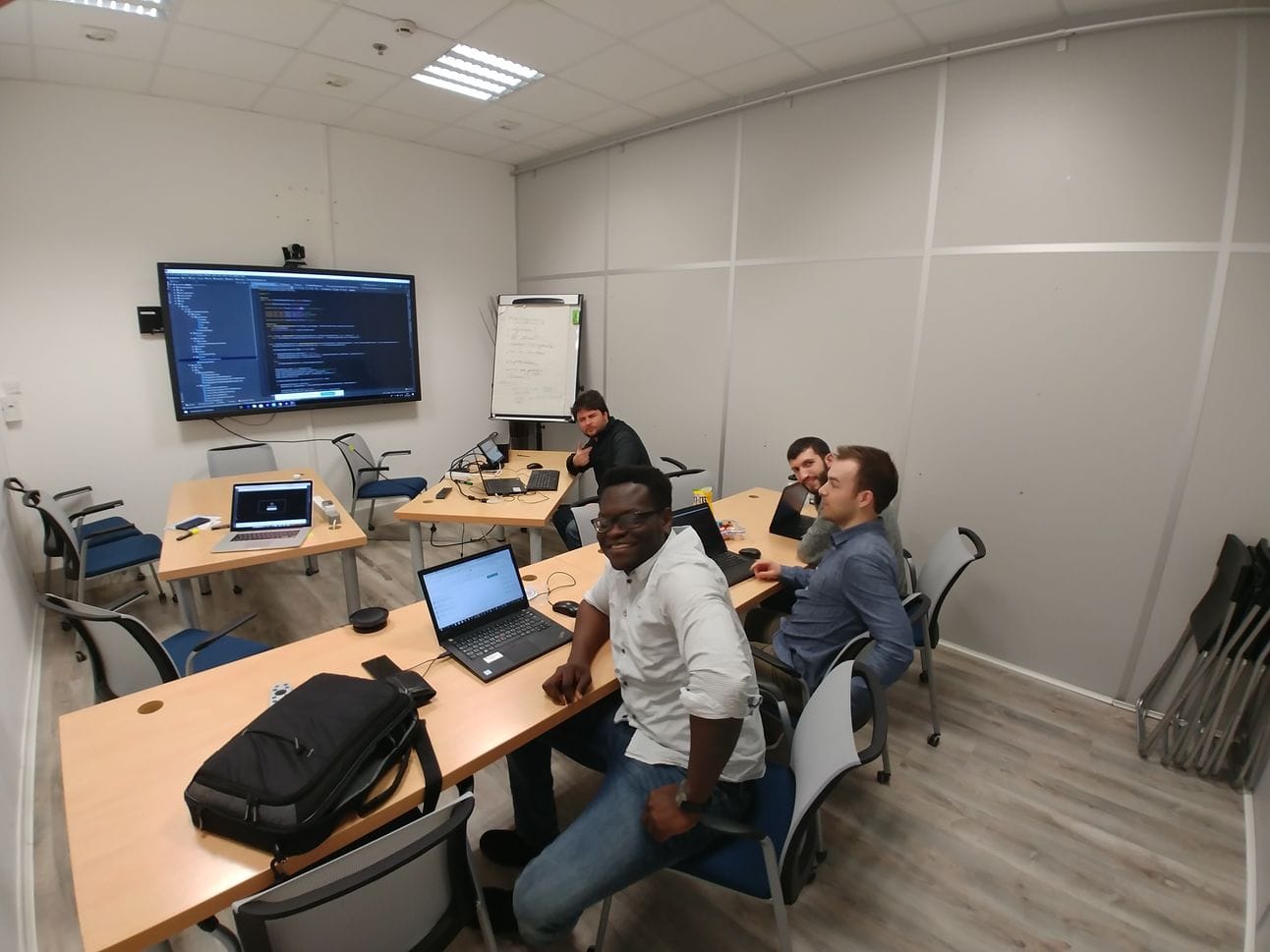 a mob programming session in a small meeting room or collaborative workspace. The room is set up with several people working together at wooden desks arranged in a row, with a large wall-mounted display showing code on the left wall. There's also a whiteboard visible with some notes.